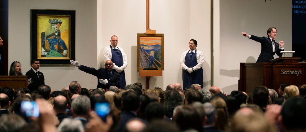 En auksjonarius leder en auksjon og peker mot et fargerikt, abstrakt kunstverk. Det er mange mennesker i salen. Foto.