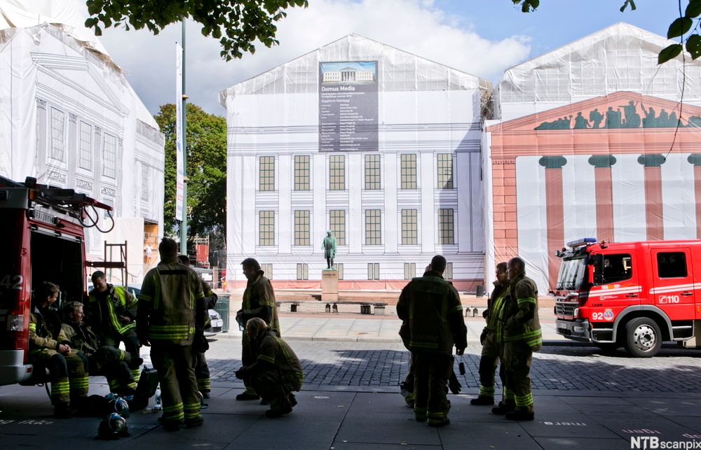 Flere personer kledd i beredskapsdrakter er ved siden av brannbiler i ei gate. De har tatt hjelmene av og sitter og står og diskuterer. Foto.