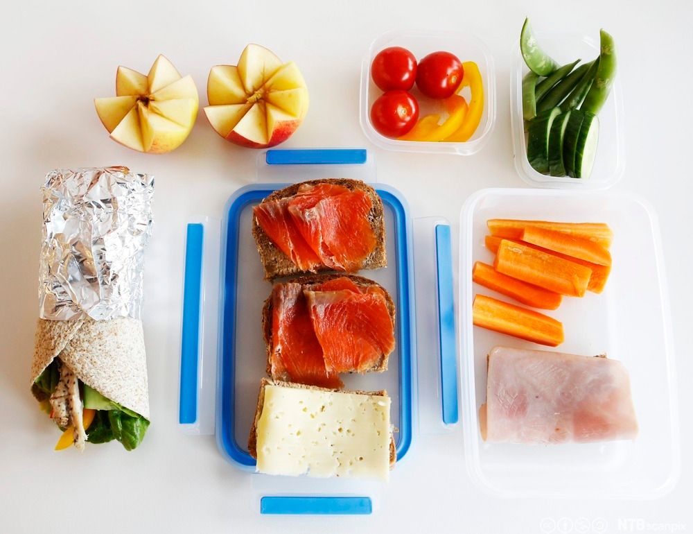 Matboks med gulrotstenger og brødskiver med ost, skinke og røkelaks. To mindre matbokser med tomater, paprika, agurk og sukkererter. Et delt eple. En sammenrullet tortillalefse med kylling og grønnsaker. Foto.