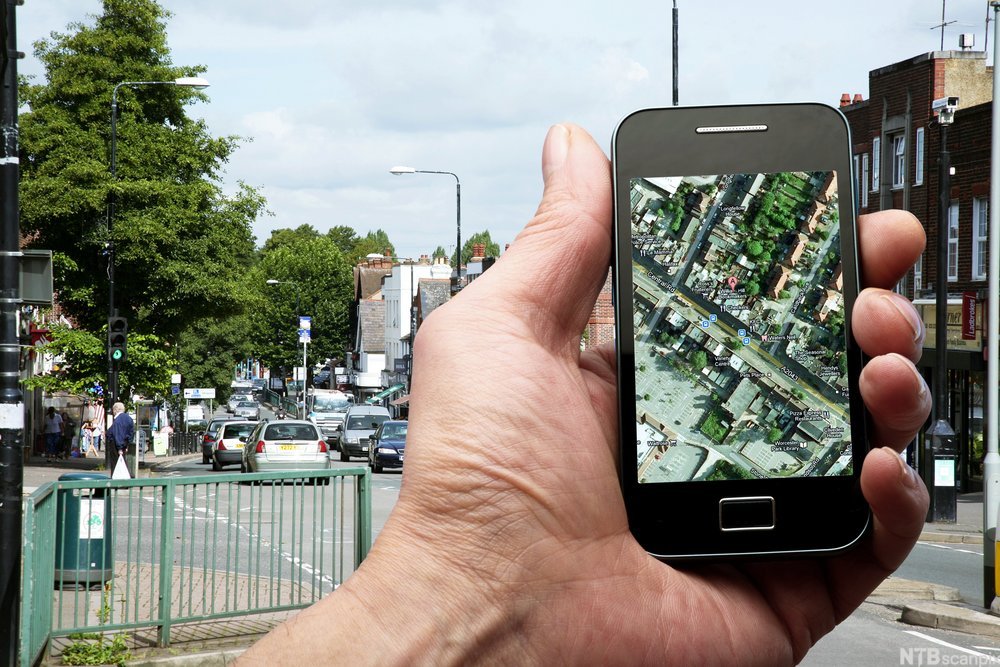 Hånd holder en smarttelefon med et satelittkart på skjermen. Bybilde i bakgrunnen. Foto.