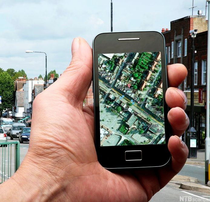 Hånd holder en smarttelefon med et satelittkart på skjermen. Bybilde i bakgrunnen. Foto.