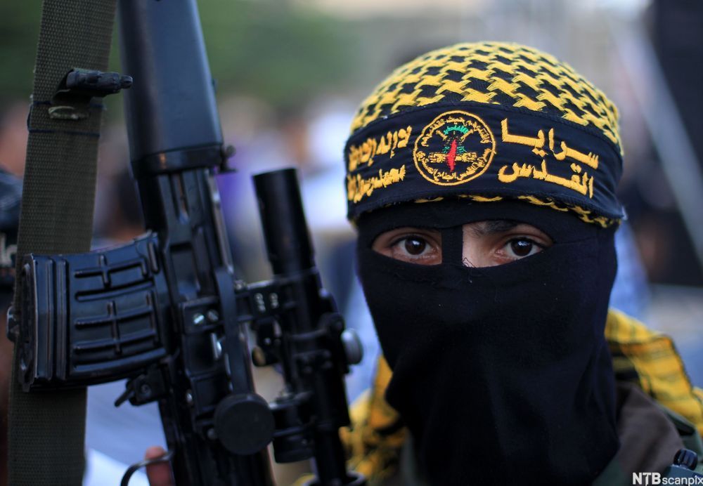 Mann med våpen og tildekt ansikt ser mot kamera. Foto.