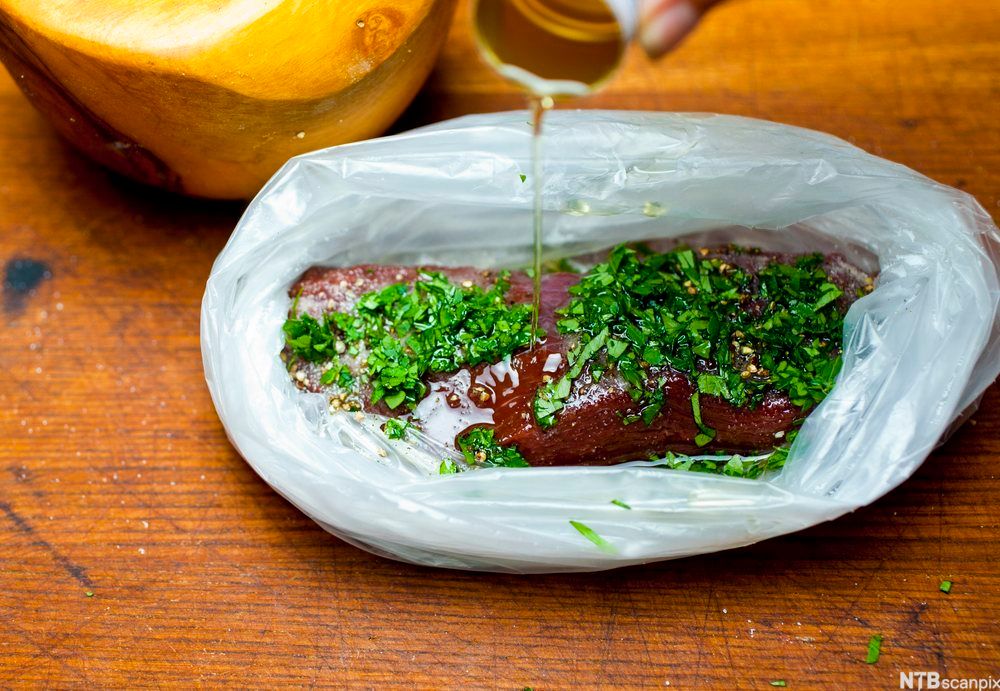 Kjøttstykke med grønne urter pakket ned i en plastpose. En væske helles over kjøttet. Foto.