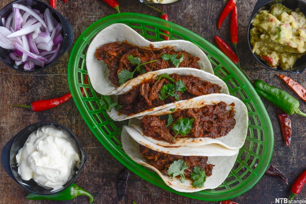 Chilitaco i ei grøn korg, guacamole, rømme og raudlauk i skåler ved sida av, saman med heile chiliar. Foto.