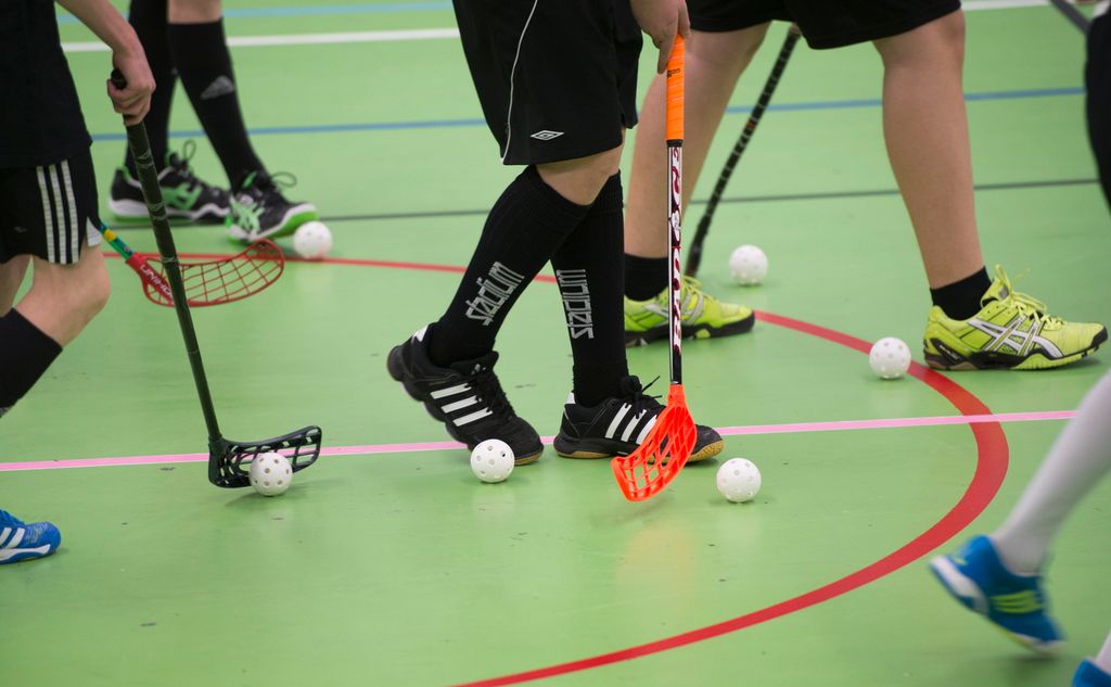 Mange innebandyspillere i en gymsal. Nærbilde av føtter, bandykøller og baller. Foto.