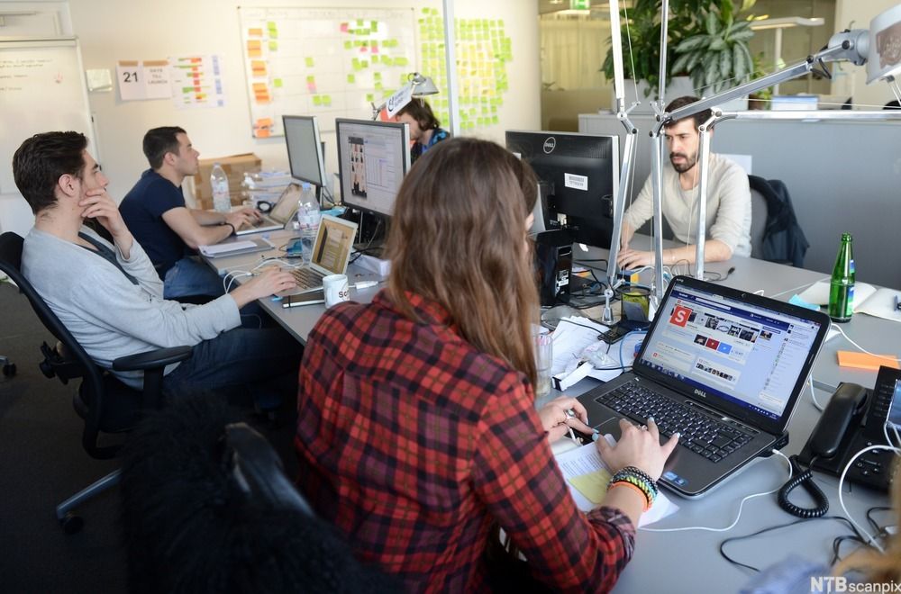 Fire personer sitter rundt et bord og jobber med datamaskiner, både bærbare og stasjonære. Foto.