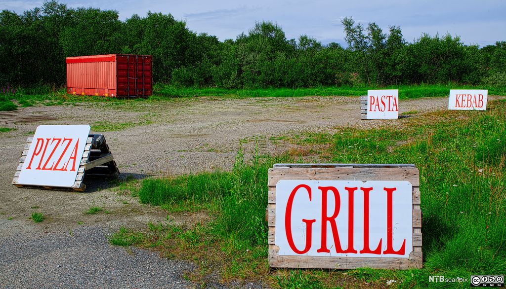 Fire store reklameplakater med ordene pizza, kebab, pasta og grill er festet til oppreiste paller. De er stilt ut på en åpen plass. Bakken er delvis dekket med grus og ugress. Inne på plassen står det også en rød container. I bakgrunnen vokser det frodig grønn vegetasjon. Foto.
