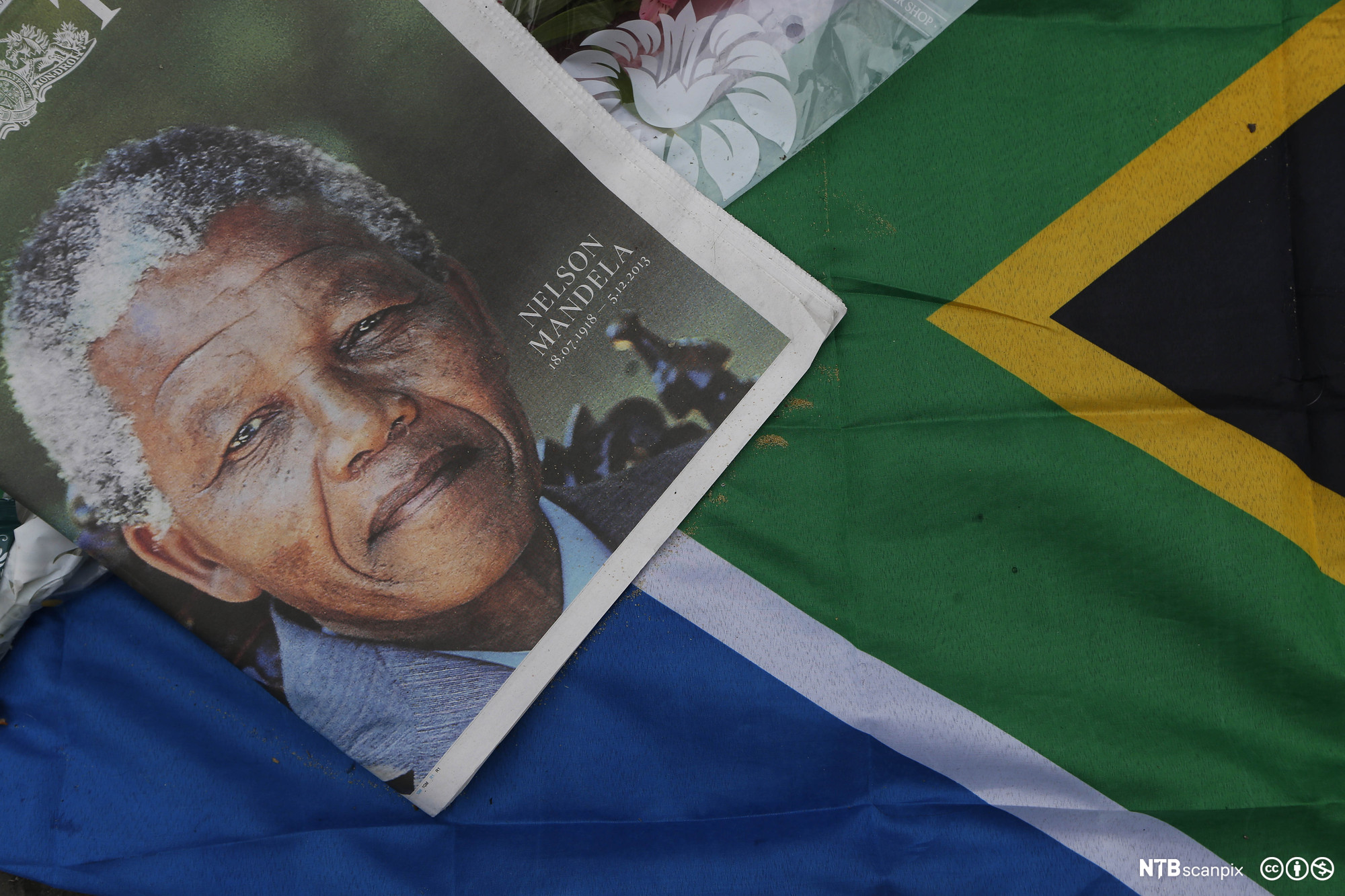En avisforside med et stort bilde av Nelson Mandela er lagt på et sørafrikansk flagg. Avisen har Mandelas navn, fødsels- og dødsdato som påskrift. Foto.