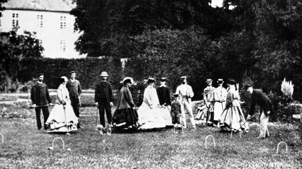 Ei gruppe staselig kledde kvinner og  menn spiller cricket i en park. Foto.