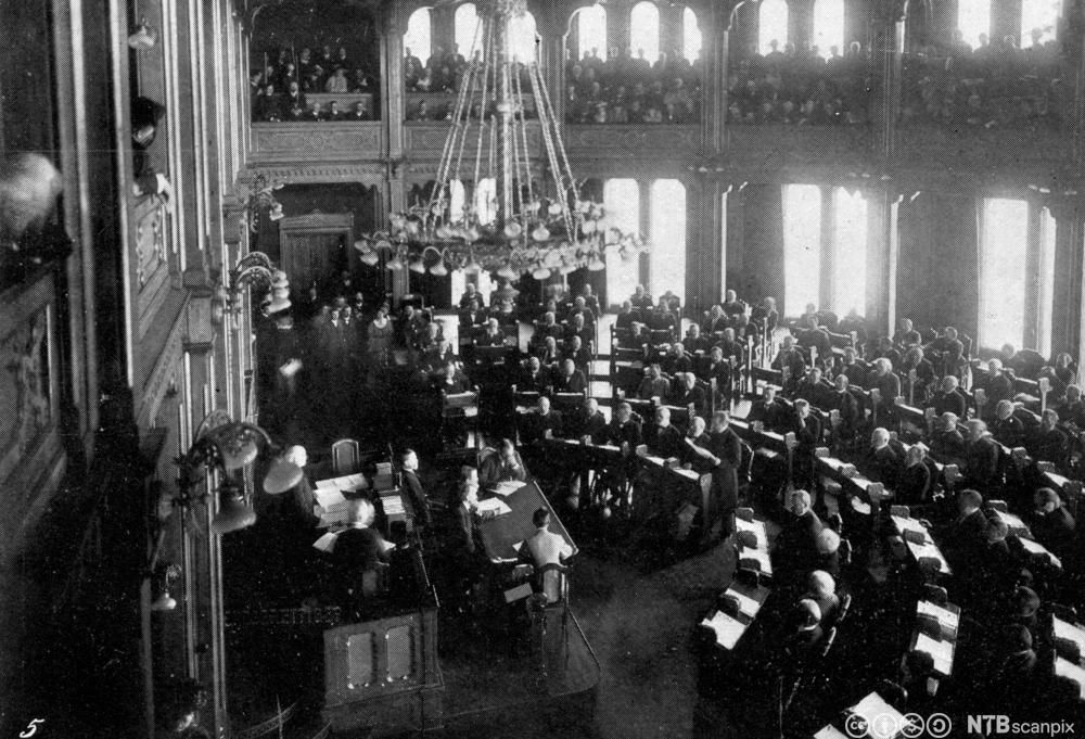 Utsyn utover en fullsatt  stortingssal. I tillegg er det fullt av tilhørere oppe på galleriene. En stor lysekrone henger ned fra taket. Svart-hvitt foto. 