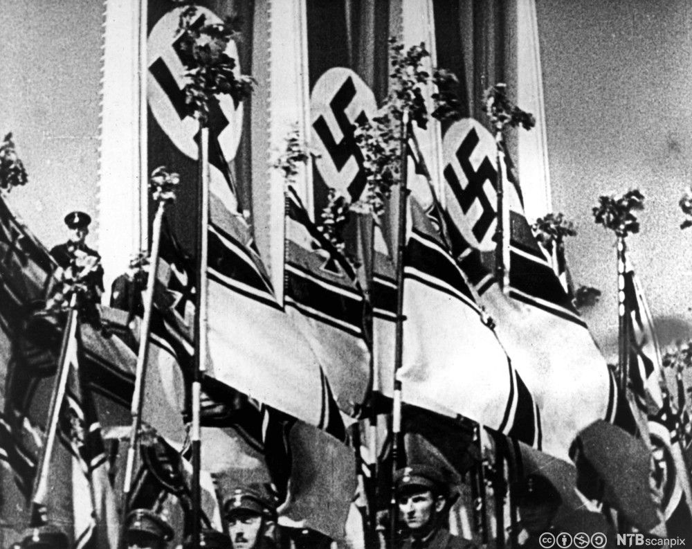 Menn i uniform marsjerer med store flagg. I bakgrunnen er det hengt opp faner med hakekors. Svart-hvitt-foto.