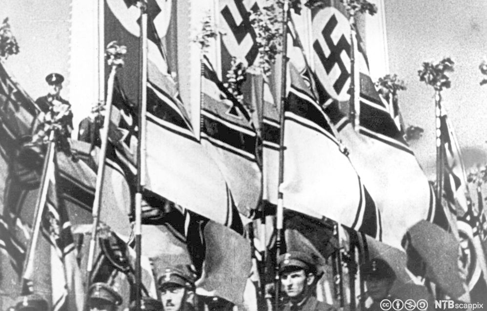 Marsjerende soldater i paradeuniform. Flagg og bannere med nazisymboler vaier over dem. Foto.