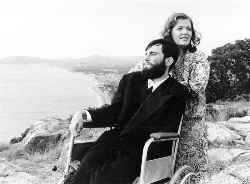 From a scene in the movie 'My Left Foot'. A man sits in a wheelchair with a woman standing behind him. They are at a lookout point. In the background you can see the sea and coastline. Photo.