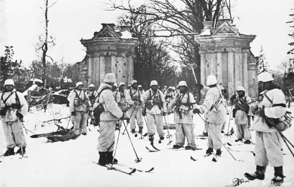 Hvitkledde soldater på ski står foran en portal eller lignende i klassisk arkitektur. I bakgrunnen en allé av trær. Foto.