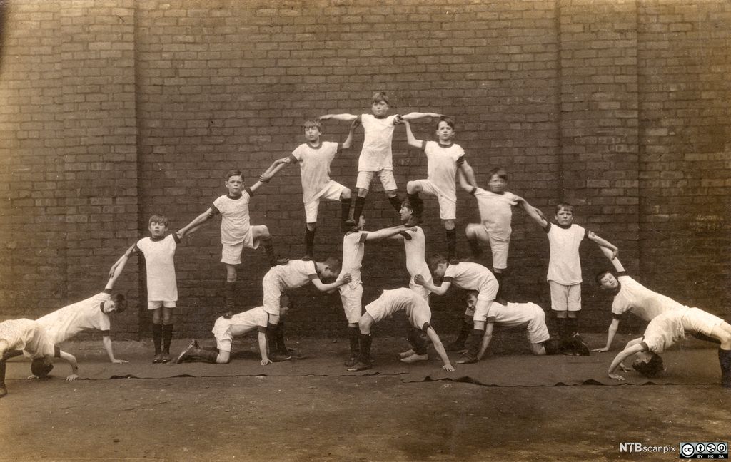 Historisk bilde av gutter som lager en menneskelig pyramide. Alle guttene har like, hvite t-skjorter og shorts og ser alvorlige ut. Foto.