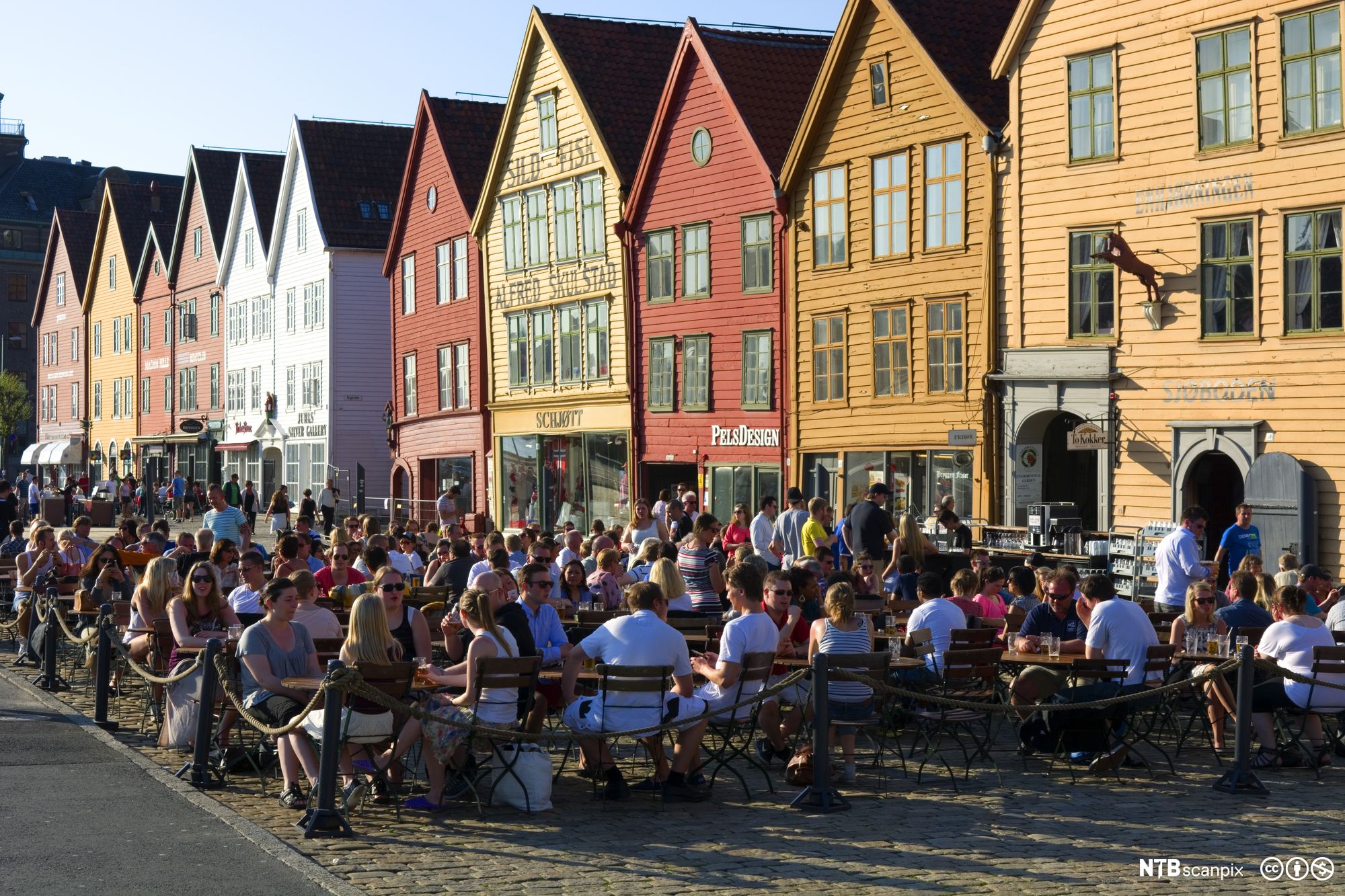 Folk sit tett i tett på uteserveringar framfor ei lang rekke trehus som er måla i raudfargar, oker eller kvitt. Foto.