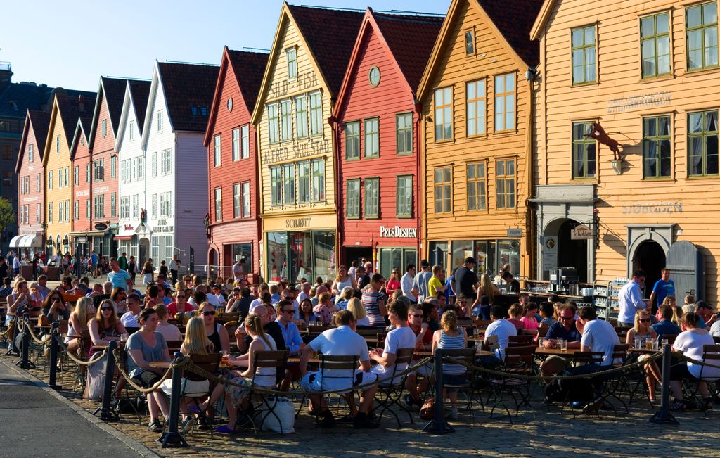 Folk sit tett i tett på uteserveringar framfor ei lang rekke trehus som er måla i raudfargar, oker eller kvitt. Foto.