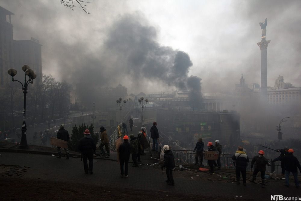 Ansamling av folk med hjelm på hodet i et bymiljø. Fra noen bygninger stiger det opp røyk som farger himmelen grå. Foto. 
