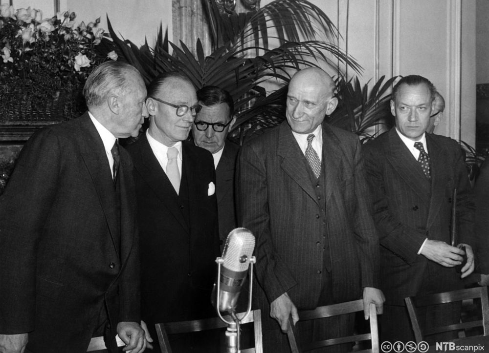 En liten gruppe menn i dress står sammen bak noen stoler. I forkant er det en mikrofon. Svart-hvitt foto. 