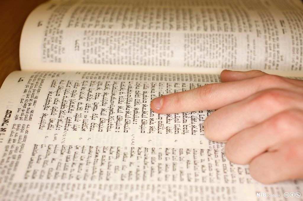 Oppslått bok med hebraisk skrift. Sidene er inndelt i ulike kolonner og tekstbokser. En hånd ligger på boken. Pekefingeren følger teksten. Foto.