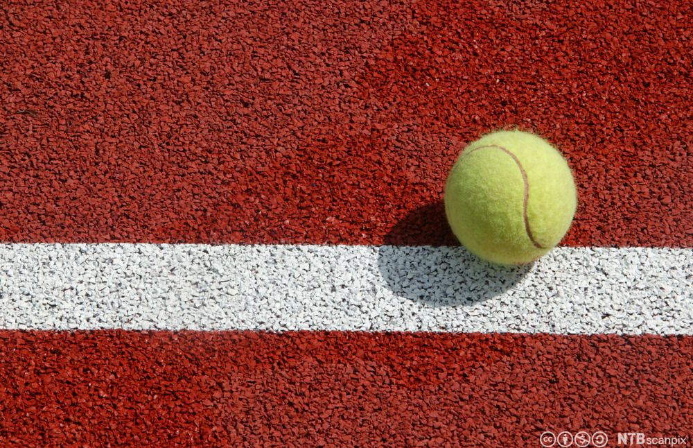 Tennisball som ligger på ei hvit linje malt på et syntetisk grusdekke. Foto.