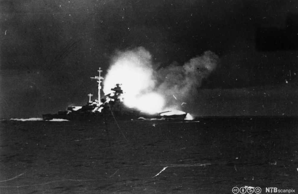 Et skip i silhuett mot røyk i bakgrunnen. Svart-hvitt foto.