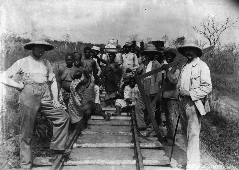 Både menn og kvinner i ulike klær står ved et jernbanespor. Foto. 