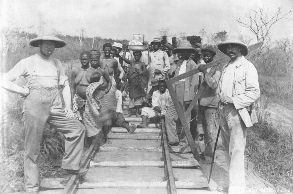 Både menn og kvinner i ulike klær står ved et jernbanespor. Foto.