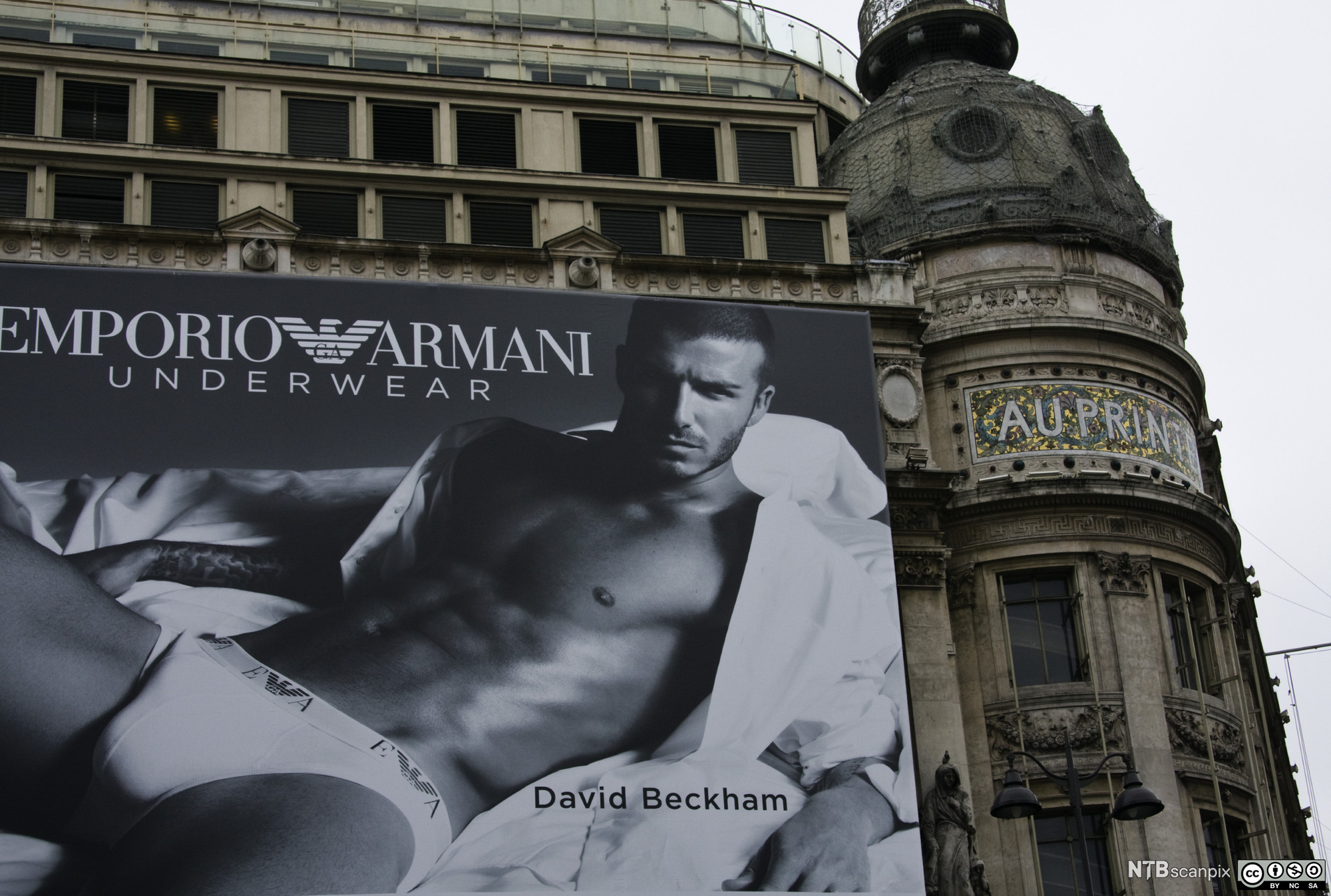 Stor plakat av en person som ligger i undertøyet og med åpen skjorte på en seng. På plakaten står det Emporio Armani Underwear. Plakaten er montert på en bygningsfasade. Foto.