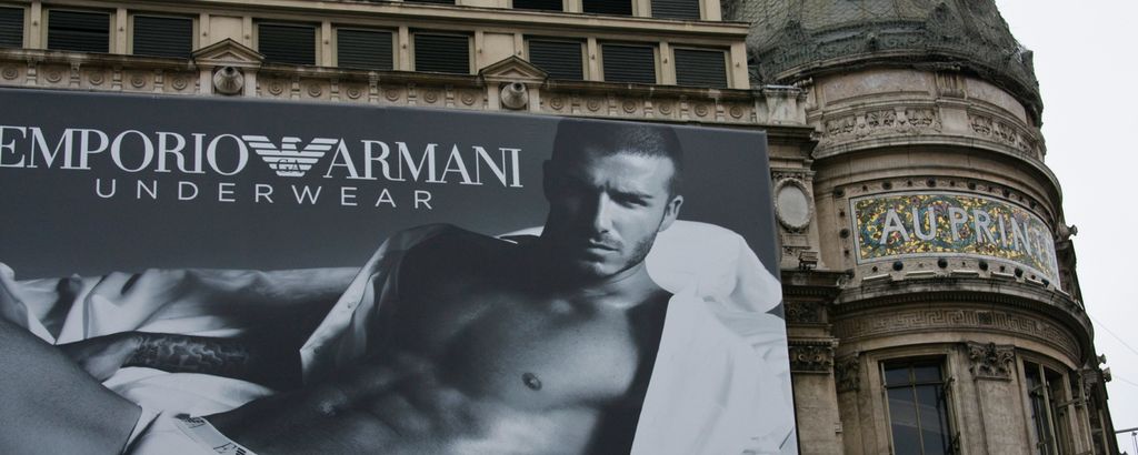Stor plakat av en person som ligger i undertøyet og med åpen skjorte på en seng. På plakaten står det Emporio Armani Underwear. Plakaten er montert på en bygningsfasade. Foto.