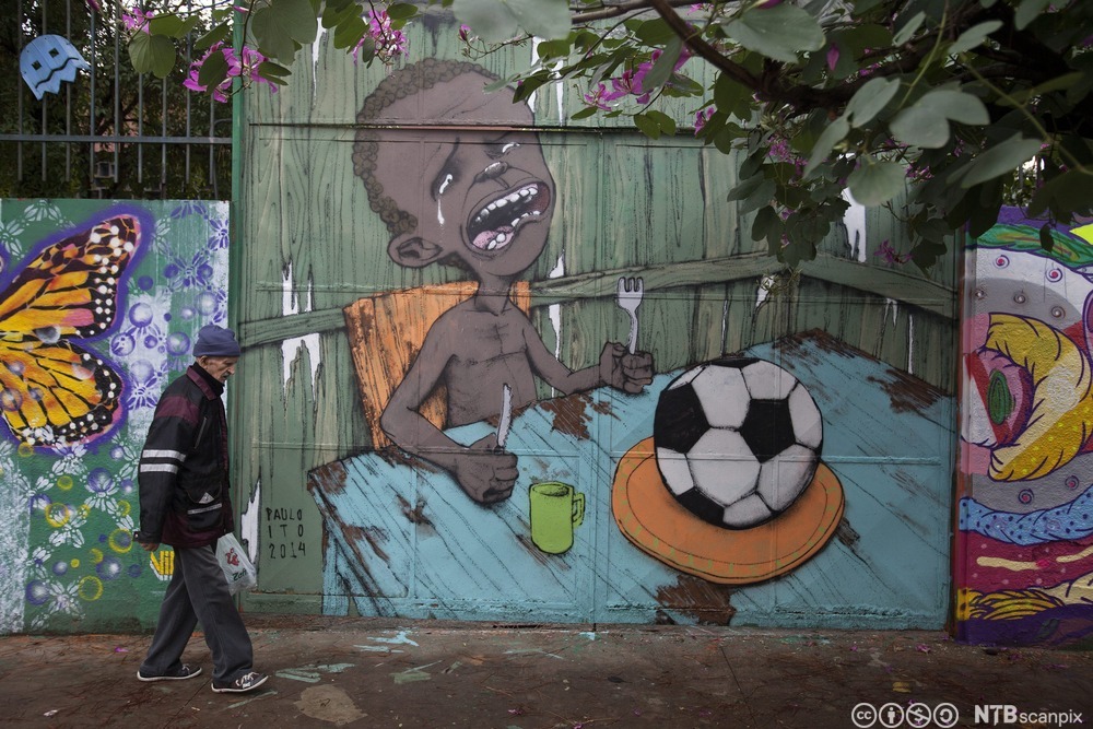 Grafitti på en vegg som viser en gråtende gutt ved et spisebord. På fatet hans ligger det en fotball. En eldre mann går forbi veggen. Foto.