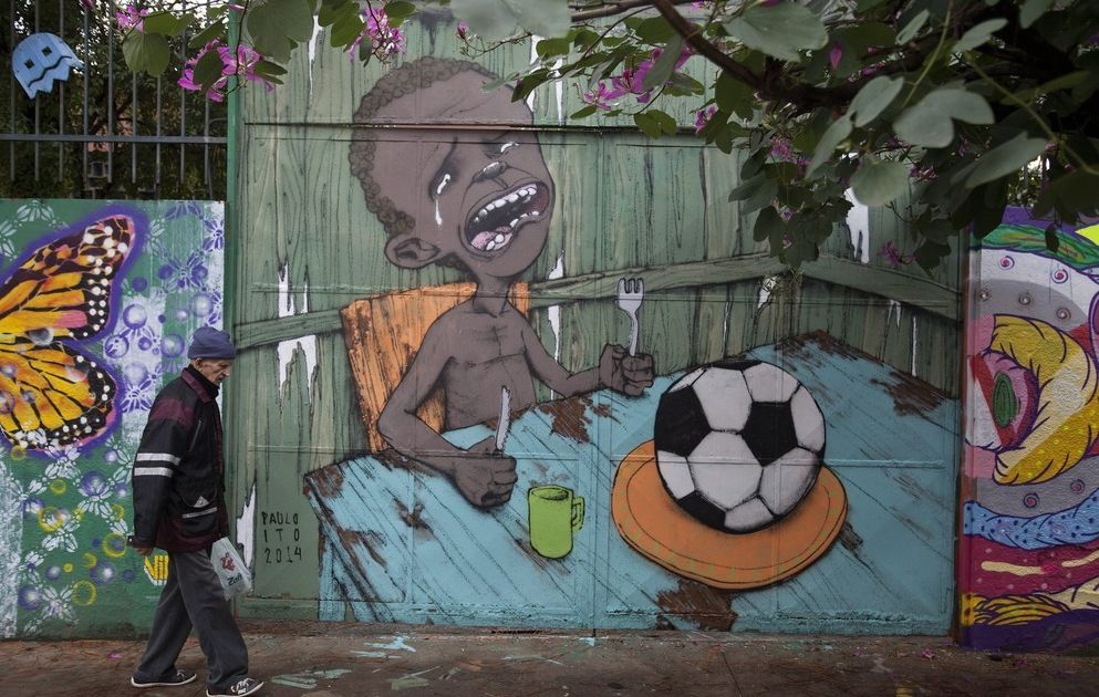Grafitti på en vegg som viser en gråtende gutt ved et spisebord. På fatet hans ligger det en fotball. En eldre mann går forbi veggen. Foto.