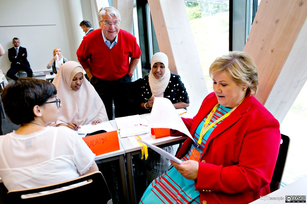 Erna Solberg sitter sammen med tre elever ved to pulter som er satt sammen. Solberg blar gjennom noen ark. Fabian Stang står mellom to av elevene og bøyer seg ned til gruppa. Foto.