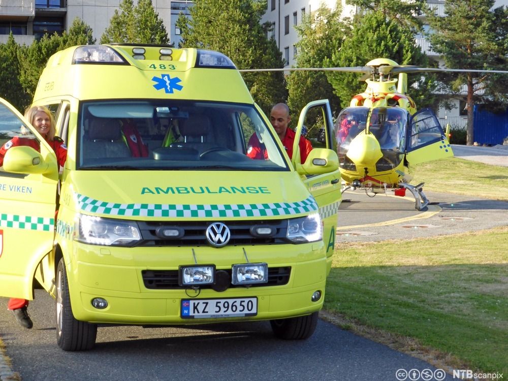 Ein gul ambulanse med opne dører står i framgrunnen. To ambulansearbeidarar i raude uniformer er på veg inn i bilen. I bakgrunnen står eit gult ambulansehelikopter på ein landingsplass. 
