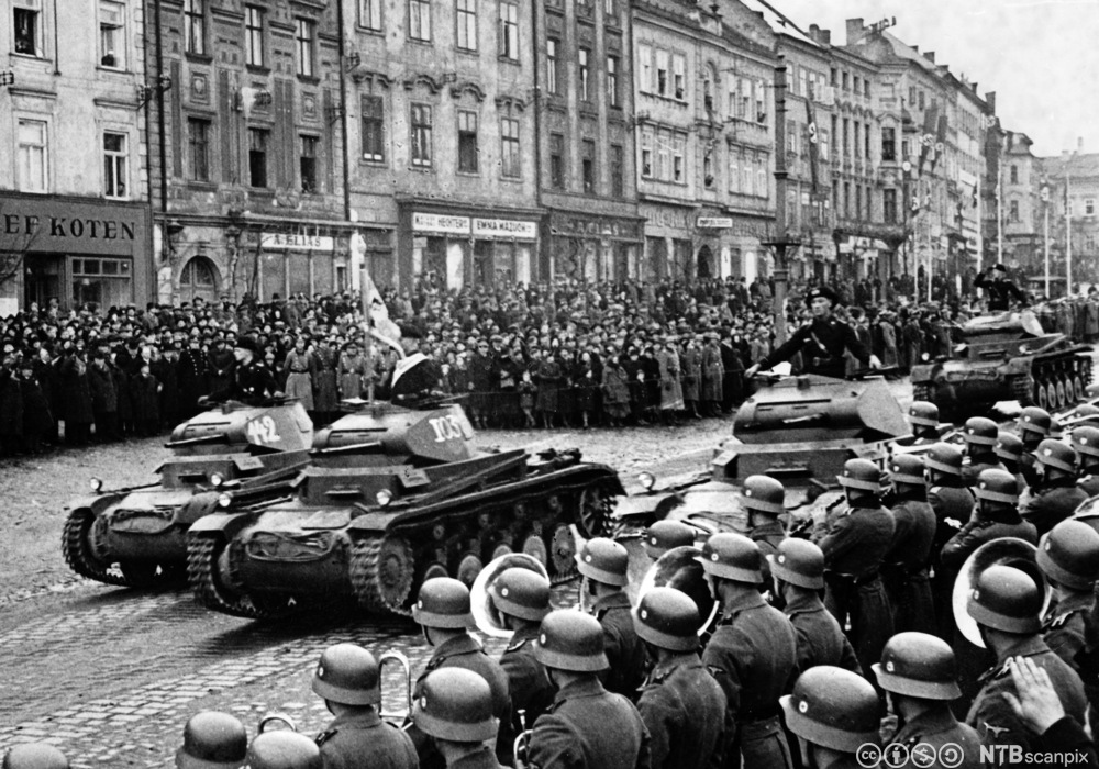 Tanks ruller inn i en by. Det står soldater med hjelmer langs den ene siden av gata i forkant av bildet. På den andre siden er det en stor folkemengde foran klassiske bygårder med forretninger i første etasje. Foto. 