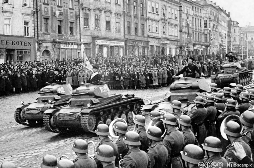 Tanks ruller inn i en by. Det står soldater med hjelmer langs den ene siden av gata i forkant av bildet. På den andre siden er det en stor folkemengde foran klassiske bygårder med forretninger i første etasje. Foto.