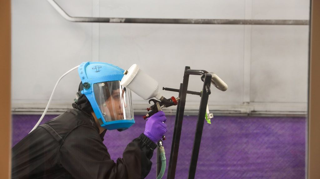 En person lakkerer et motorsykkelstyre med ei luftsprøyte. Personen har verneklær og lufttilført vernemaske. Foto.