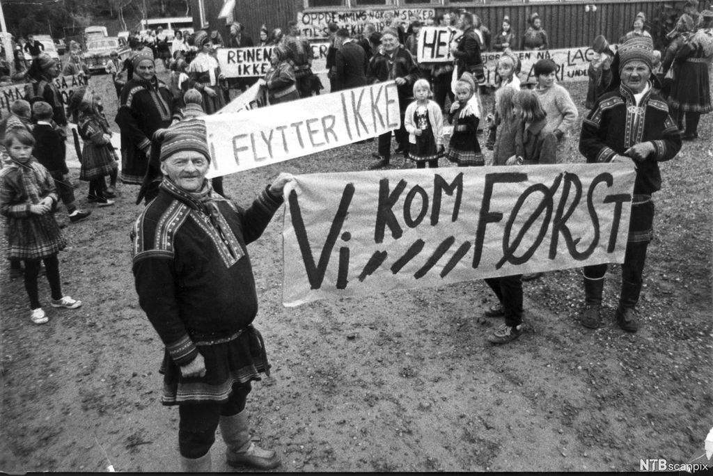 Menn, kvinner og barn, noen av dem ikledd samiske drakter, med bannere der det står "Vi kom først" og "Vi flytter ikke". En bygning og en parkeringsplass i bakgrunnen. Foto.