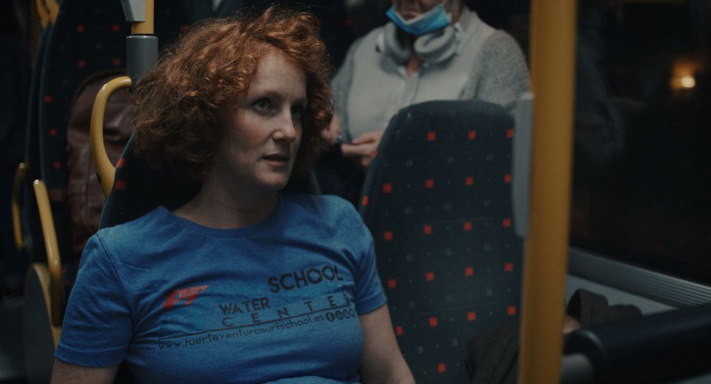 En ung kvinne med rødt hår og blå genser sitter på en buss. Hun har et skeptisk uttrykk. Foto.