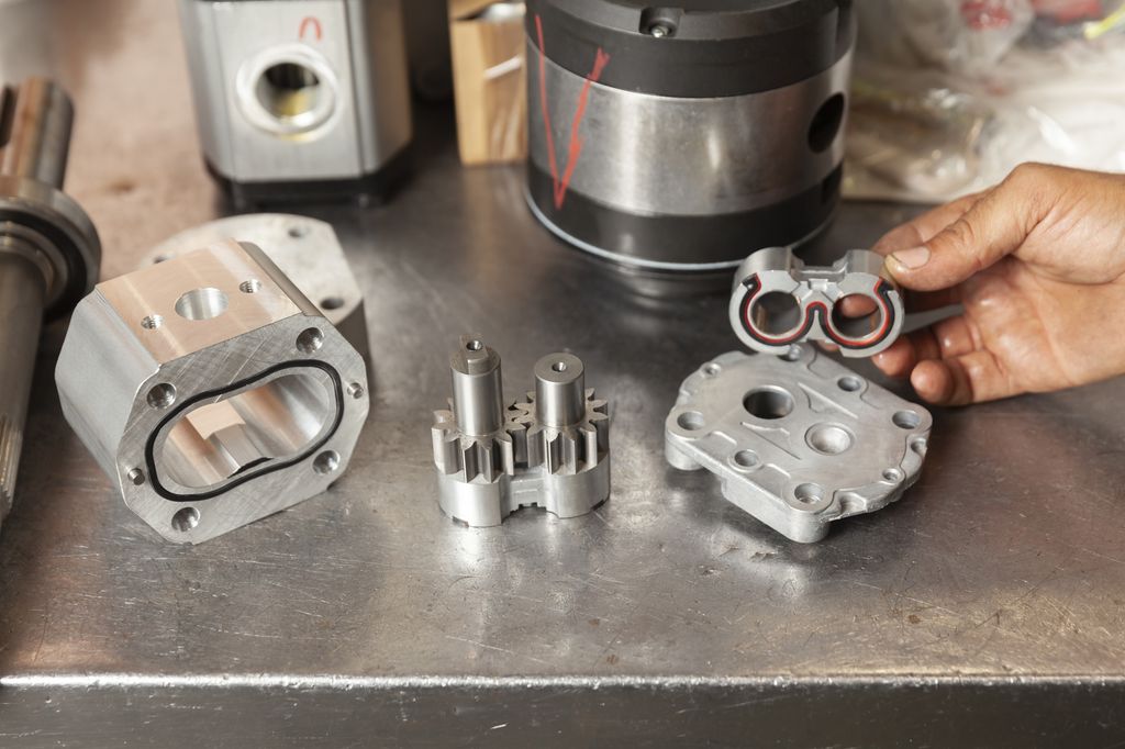 Reparasjon av hydraulisk pumpe på verkstad. Pumpa er demontert, det ligg tre delar på eit bord, og ein person held den siste delen. Foto.