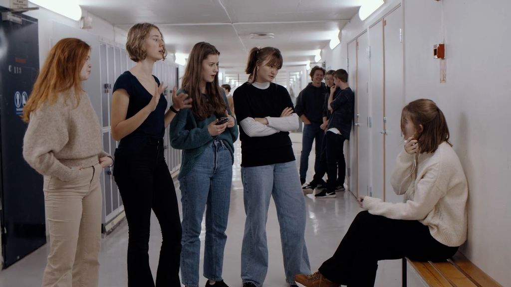 Fire jenter står overfor ei annen jente som sitter på en benk i en skolekorridor. Foto