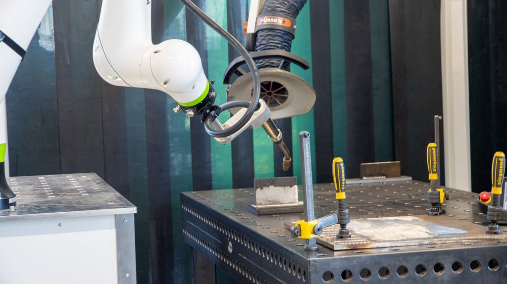 En robot med en sveisepistol stående ved et metallbord med ulike verktøy og metallplater. Foto.