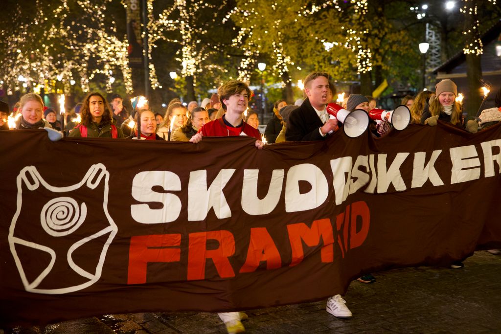 Ungdommer marsjerer bak et banner med bilde av en skuddsikker vest og teksten "Skuddsikker framtid" på. Foto.