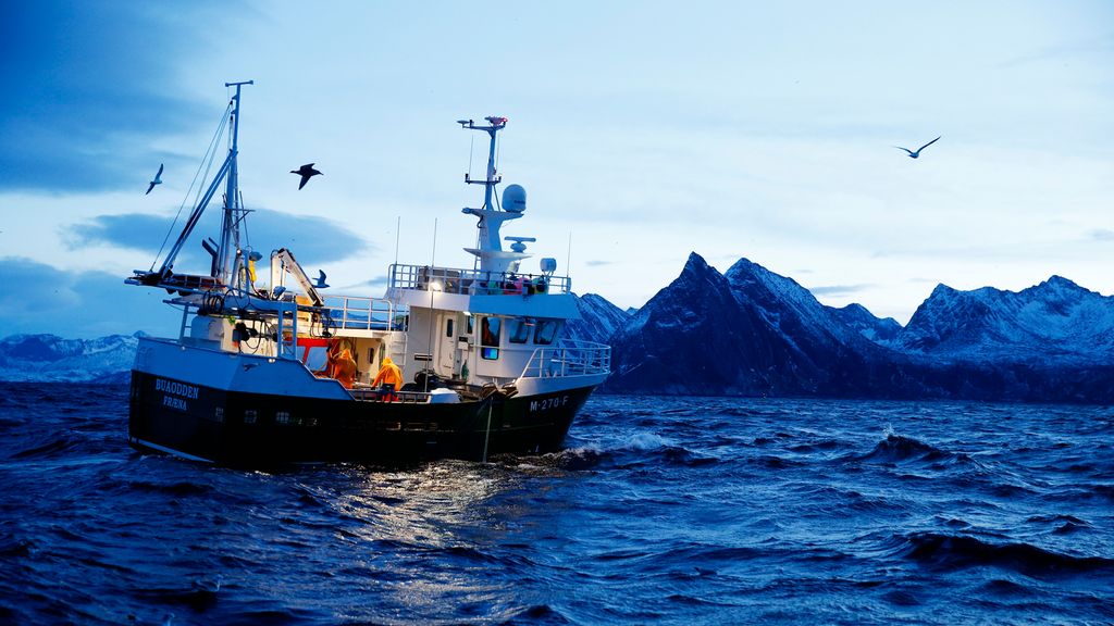 Fiskebåt på havet. Spisse fjell i bakgrunnen. Foto.