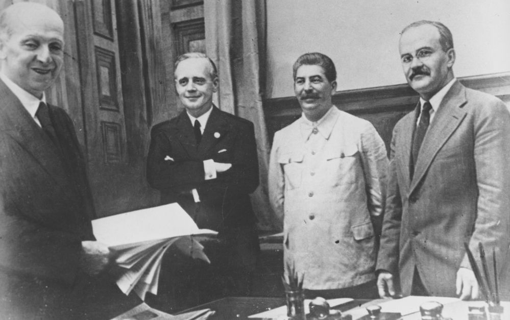 Fire smilende, middelaldrende menn bak et arbeidsbord. En bunke papirer holdes fram. Stalin er litt annerledes kledd med en uniformslignende jakke. Foto.