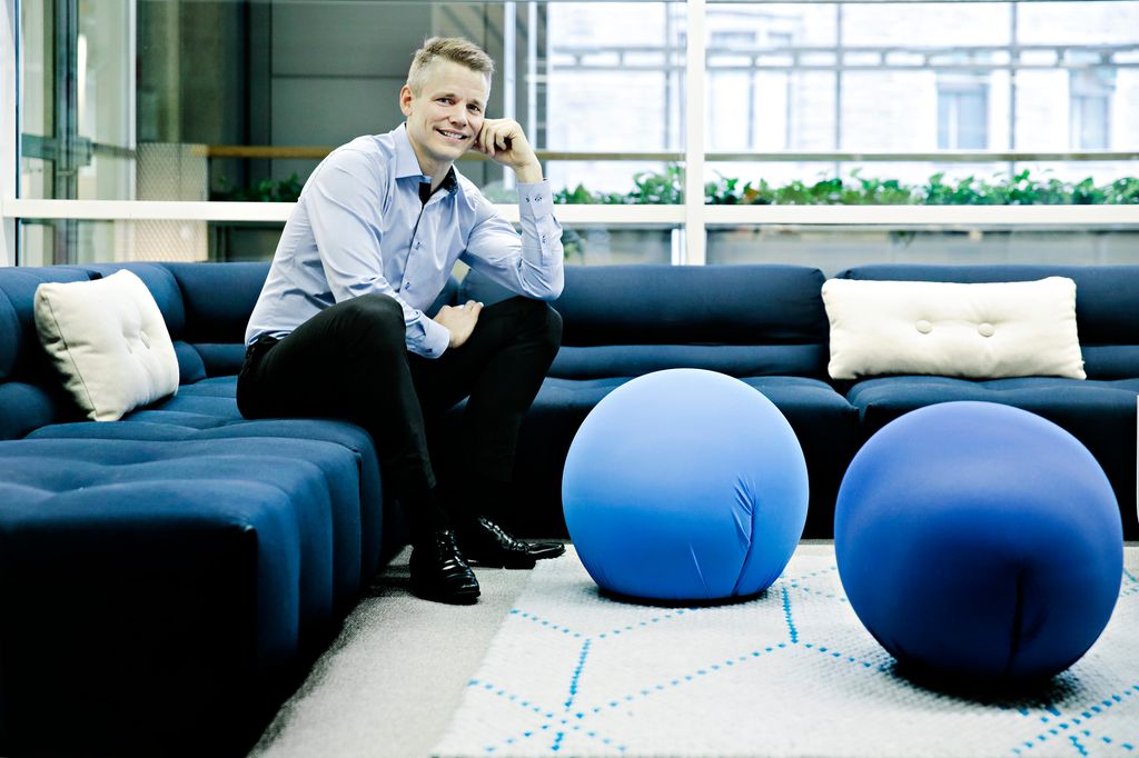 En mann sitter i en blå sofa i et kontorlandskap. Foto.
