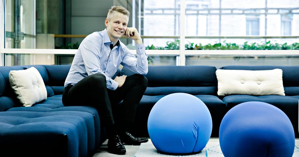 Ung mann i skjorte og dressbukse sitter i en stor blå sofa i et kontorlandskap og smiler mot fotografen. Foto.