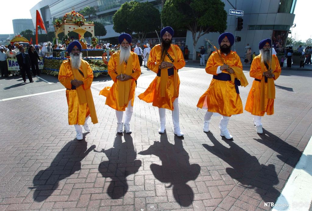 Fem menn med sverd i orange klær og blå turban går først i en parade. Foto.