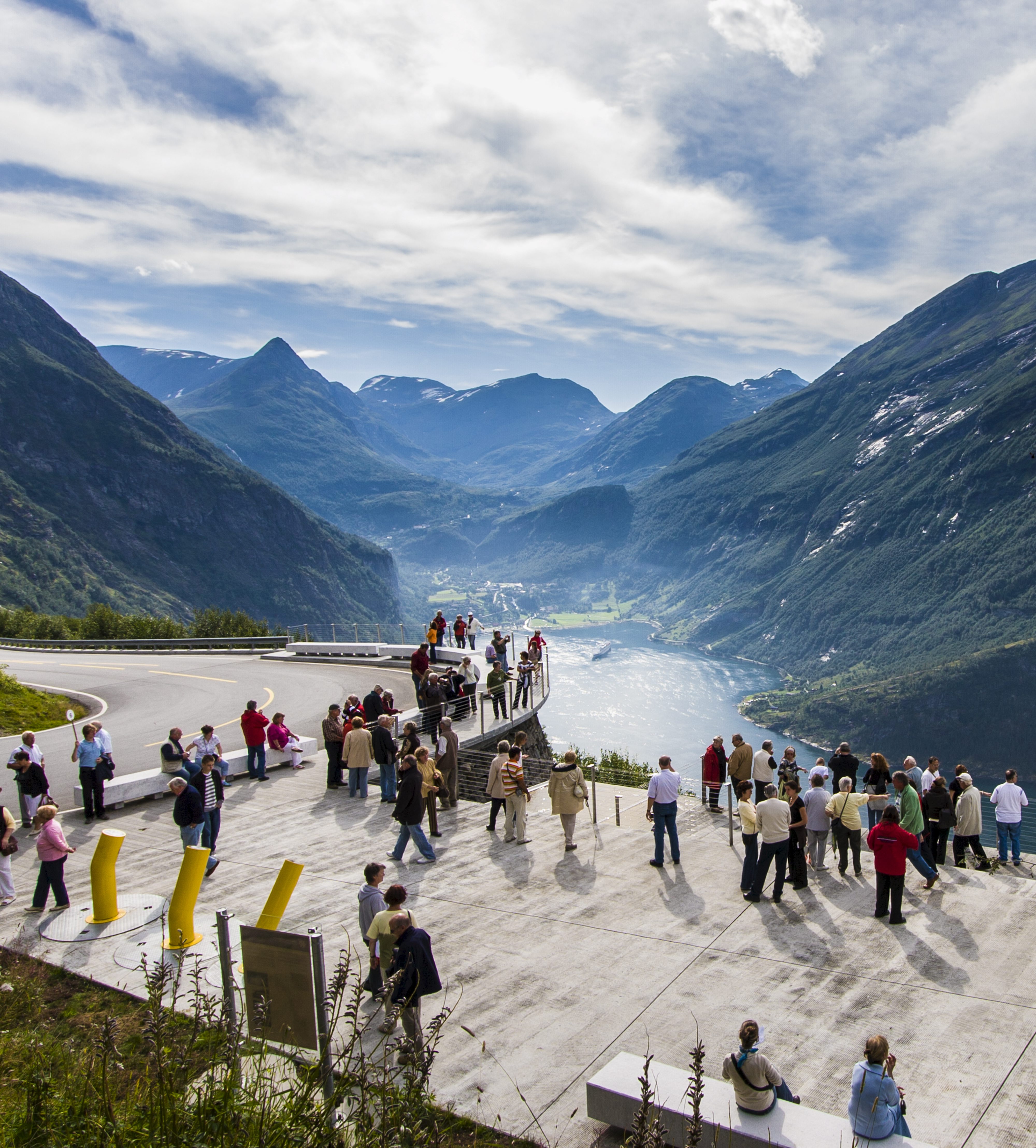 Turister skuer utover et fjord- og fjellandskap fra et utkikkspunkt langs en bilvei. Foto.