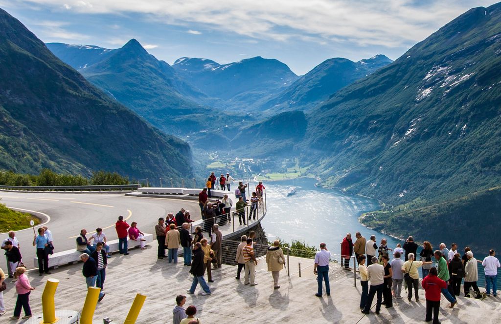 Turister skuer utover et fjord- og fjellandskap fra et utkikkspunkt langs en bilvei. Foto.
